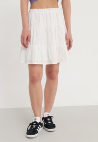 Femme portant une mini-jupe blanche à volants et des baskets Adidas noires, appuyée contre un fond blanc neutre.