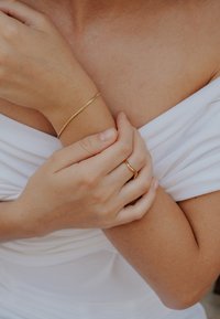 Zlatý náramok a prsteň na ruke; náramok má hladký, minimalistický dizajn. Tón pokožky je teplý, s jemnou látkou splývajúcou v pozadí.