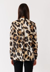 Camicia a maniche lunghe in tessuto setoso con stampa leopardata beige e nera, colletto casual e polsini con bottone. Sono visibili dei pantaloni neri.