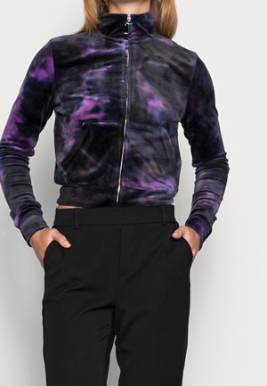 Kobieta ubrana w fioletowo-czarną kurtkę z nadrukiem tie-dye zapinaną na suwak, z rękami w kieszeniach wysokich czarnych spodni, na jednolitym tle.