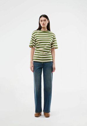 Kobieta stojąca, ubrana w zielono-beżowy pasiasty t-shirt, niebieskie jeansy o prostych nogawkach i brązowe buty na białym tle.