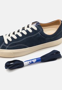 Sapatilha em camurça azul-marinho com atacadores creme e sola de borracha, apresentando costuras brancas em contraste e um rótulo com o logótipo na língua. Inclui atacadores extras em azul-marinho.