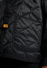 Musta tikattu takki, jossa on teksturoitu, pehmustettu pinta, näkyvä läppätasku ja oranssia kontrastivärinä oleva logotunniste. Edessä mustat napit.