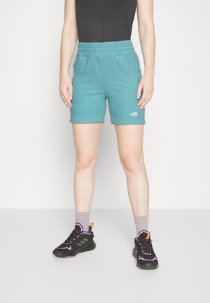 Persoon die tealgekleurde The North Face shorts draagt, een zwart topje, grijze sokken en zwarte sneakers met roze en oranje accenten, staand op een lichte vloer.