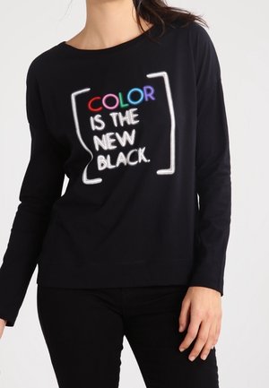 Osoba ubrana w czarną koszulę z długim rękawem z napisem „COLOR is the new BLACK” w białych i wielokolorowych literach na przodzie.