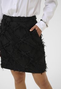 Černá sukně s texturovaným designem, s třepícími se detaily a diamantovým vzorem. Obsahuje kapsu, v kombinaci s bílou košilí na knoflíky.