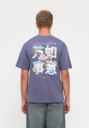 Young man facing away wearing blue T-shirt with floral design and text "Good luck in everything" and loose denim jeans.