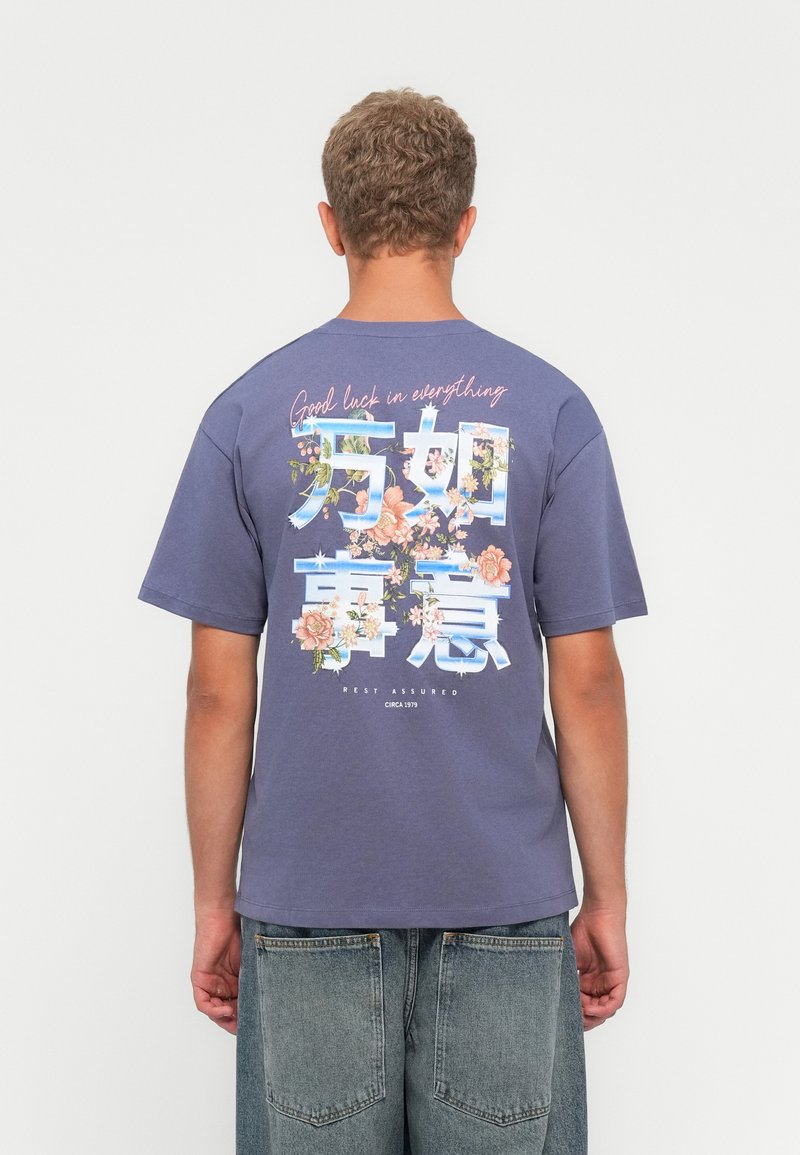 Joven de espaldas con una camiseta azul de diseño floral y el texto "Buena suerte en todo" y jeans de mezclilla sueltos.