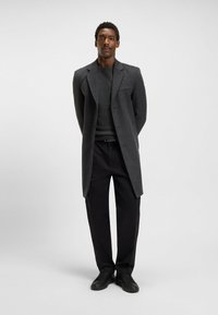 Cappotto grigio di lana con vestibilità sartoriale, design monopetto e colletto a punta. Abbinato a un maglione scuro e pantaloni neri, completato con scarpe nere.