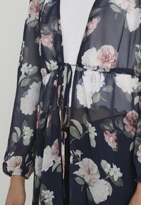 Průhledný námořnický kimonový plášť s růžovými a bílými květinami, svázaný v pase přes bílý texturovaný top.