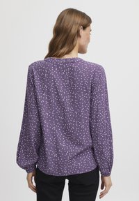 Lila långärmad blus med vita prikar, mjukt tyg, rundad nederkant och samlade manschetter; bakvy visar en avslappnad passform och krage.