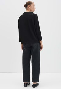 Frau mit hochgestecktem Haar, die einen lockeren schwarzen Pullover, weite, ausgebleichte schwarze Jeans und schwarze Slipper trägt, mit dem Rücken zu einer schlichten Hintergrundwand.