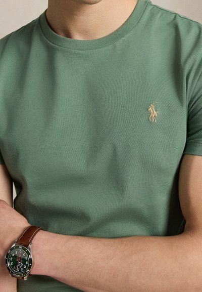 Moški, ki nosi zeleno majico Polo Ralph Lauren z bež logotipom in uro z rjavim usnjenim paščkom na levi zapestju, rokami prekrižanimi v višini pasu.