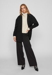 Črna oversize jakna z dvema žepoma na prsih, krema pulover s turtleneck pletenjem, široke črne hlače in črne pete čevlje.