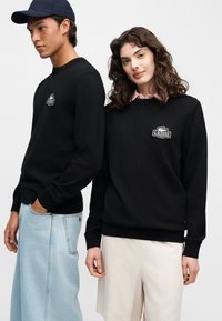 Мъж и жена носят черни суитшърти Lacoste; мъжът е с светлосини дънки и тъмносиня шапка, жената е с бял панталон и розова риза с якичка.