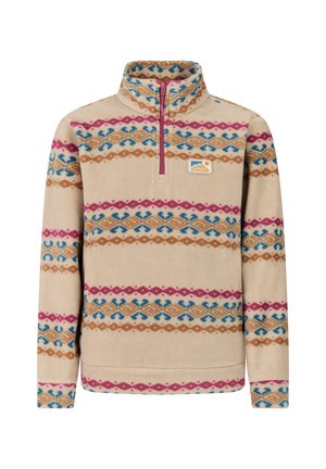 Jonge vrouw staand, draagt een beige fleece met patroon, blauwe broek en zwarte laarzen tegen een effen lichte achtergrond.