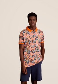 Korte mouwen polo met bloemmotief in oranje, met groene en blauwe bladeren. Het heeft een oranje kraag en manchetten, gecombineerd met een marineblauwe korte broek.