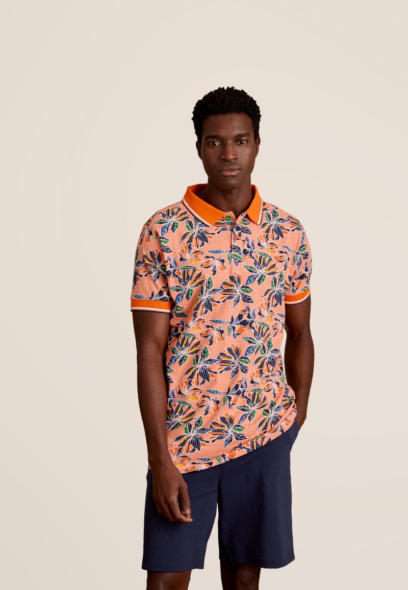 Korte mouwen polo met bloemmotief in oranje, met groene en blauwe bladeren. Het heeft een oranje kraag en manchetten, gecombineerd met een marineblauwe korte broek.
