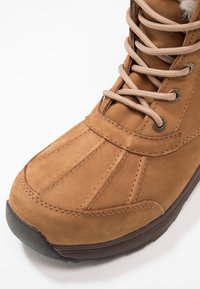 Botte de randonnée en cuir marron avec des lacets beige, des coutures détaillées et une semelle en caoutchouc foncé sur fond blanc.