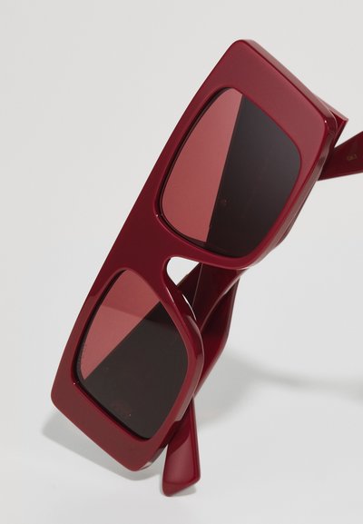 Ochelari de soare roșii de mari dimensiuni, cu rame din plastic gros și lentile întunecate. Au decupaje geometrice aproape de balamale pentru un detaliu de design.