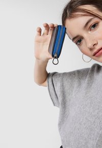 Sacca con zip realizzata in tessuto con accenti blu e una zip argentata. Forma rettangolare compatta tenuta in mano. Sfondo grigio uniforme.