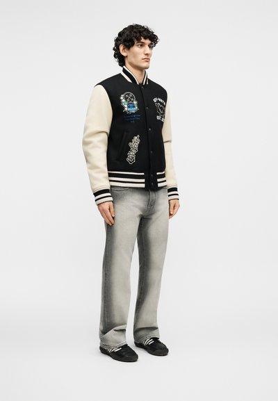 Άνδρας στέκεται φορώντας μαύρο και κρεμ varsity μπουφάν με μπαλώματα, ανοιχτό γκρι τζιν και μαύρα αθλητικά παπούτσια με λευκές ρίγες.