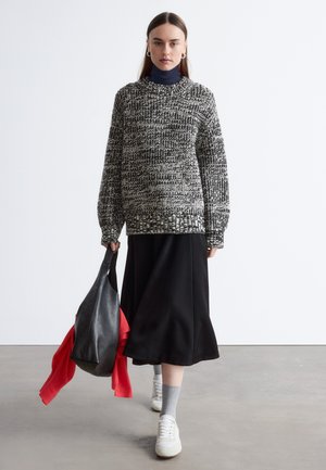 Schwarz-weiß gestrickter Pullover mit langen Ärmeln und hohem Kragen; schwarzer Rock; graue Socken; weiße Turnschuhe; schwarze Tasche mit rotem Gegenstand.