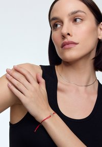 Czerwona bransoletka z regulowanym sznurkiem, z prostym metalowym charmsowym zawieszeniem. Model nosi czarny top na ramiączkach i srebrny naszyjnik choker.