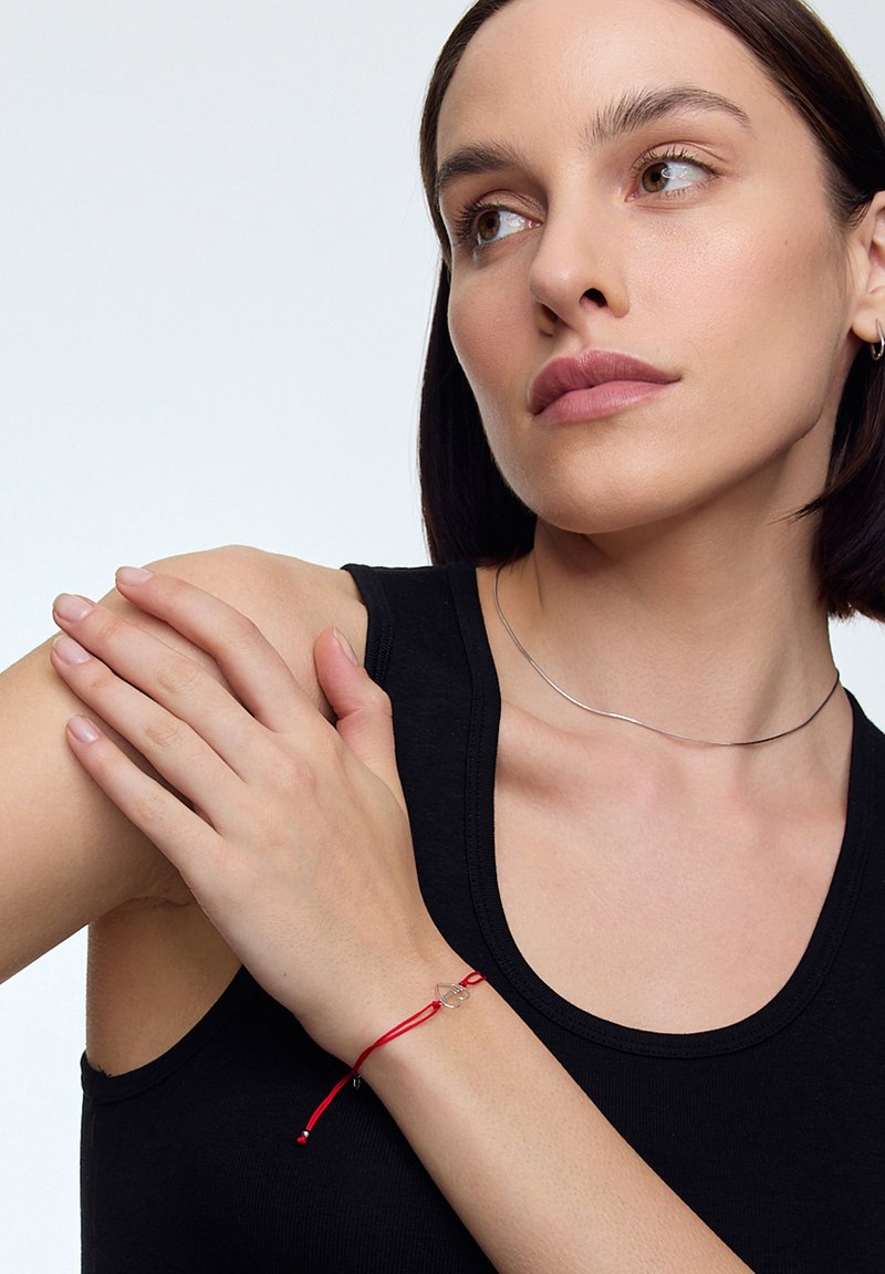 Czerwona bransoletka z regulowanym sznurkiem, z prostym metalowym charmsowym zawieszeniem. Model nosi czarny top na ramiączkach i srebrny naszyjnik choker.