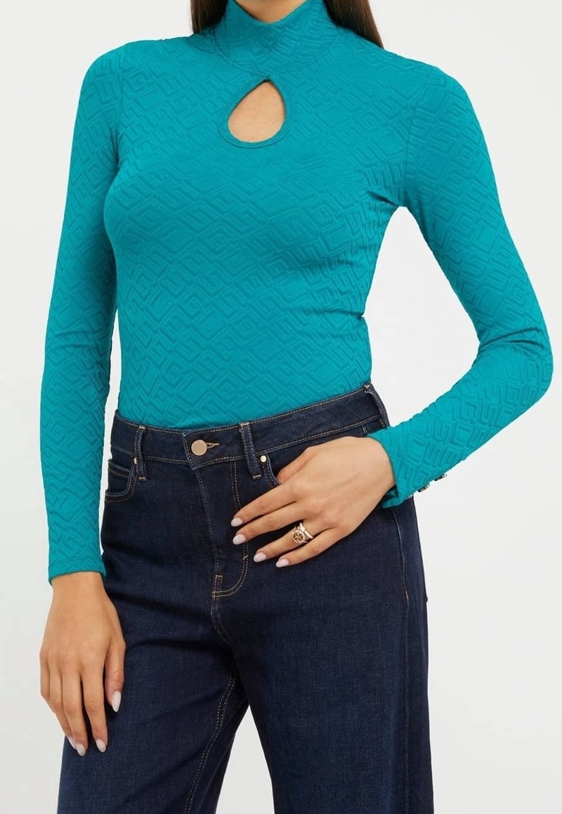 Maglia a maniche lunghe turchese con collo a mock e dettaglio a occhiello, caratterizzata da un motivo geometrico testurizzato. Abbinata a jeans in denim scuro.