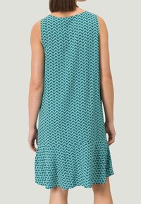 En ærmeløs, knælang kjole i teal med et bølget mønster. Den har en udvidet kant og et blødt, tekstureret stof.