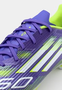 Paarse voetbalschoen met geel-groene accenten, voorzien van een strak ontwerp, een gestructureerde bovenkant en reflecterende witte strepen aan de zijkant.