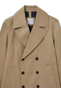 Trench coat beige doppiopetto con larghi risvolti, bottoni neri, tasche laterali e un'etichetta visibile all'interno del colletto contrassegnata "CALLIOPE."