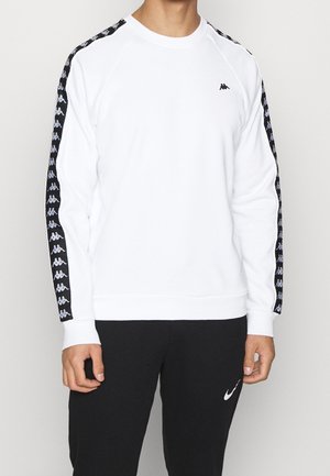Mand iført hvid sweatshirt med sort logo tape på ærmerne og sorte bukser med hvidt Nike-logo, stående foran en ensfarvet baggrund.