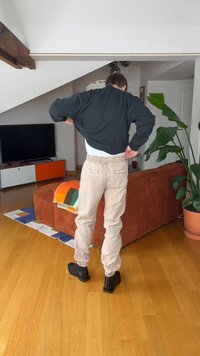 Egy férfi bézs nadrágot és fekete csizmát visel, fekete pulóvert igazítgat, miközben egy nappaliban áll, ahol barna kanapé, cserepes növény és TV-konzol található.