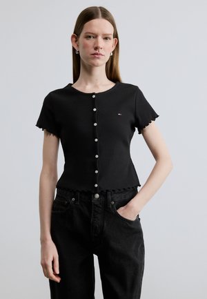 Ung kvinde med glat hår iført en sort kortærmet knapbluse og sorte jeans, står med den ene hånd i lommen.