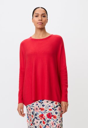 Kvinde iført en løs rød langærmet top og et cremefarvet skørt med et farverigt rødt og blåt blomstermønster, stående mod en ensfarvet baggrund.