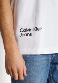 Biały bawełniany t-shirt z czarnym napisem „Calvin Klein Jeans” u dołu. Koszulka ma luźny krój i gładką teksturę.