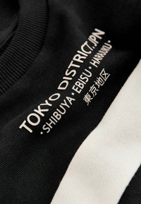 Czarna bluza z białymi poziomymi paskami, z nadrukowanym tekstem "TOKYO DISTRICT.JPN - SHIBUYA - EBISU - HARUMI" w nowoczesnym kroju.