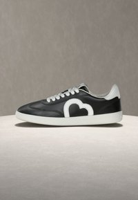 Μαύρο χαμηλό αθλητικό παπούτσι με λευκές κορδόνια, λευκή σόλα και λευκό σχέδιο σε σχήμα καρδιάς στο πλάι, τοποθετημένο σε μπεζ επιφάνεια.