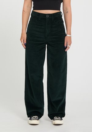 Personne portant un pantalon large en velours côtelé vert foncé taille haute, des baskets noires et blanches et un top court noir, debout devant un fond uni.