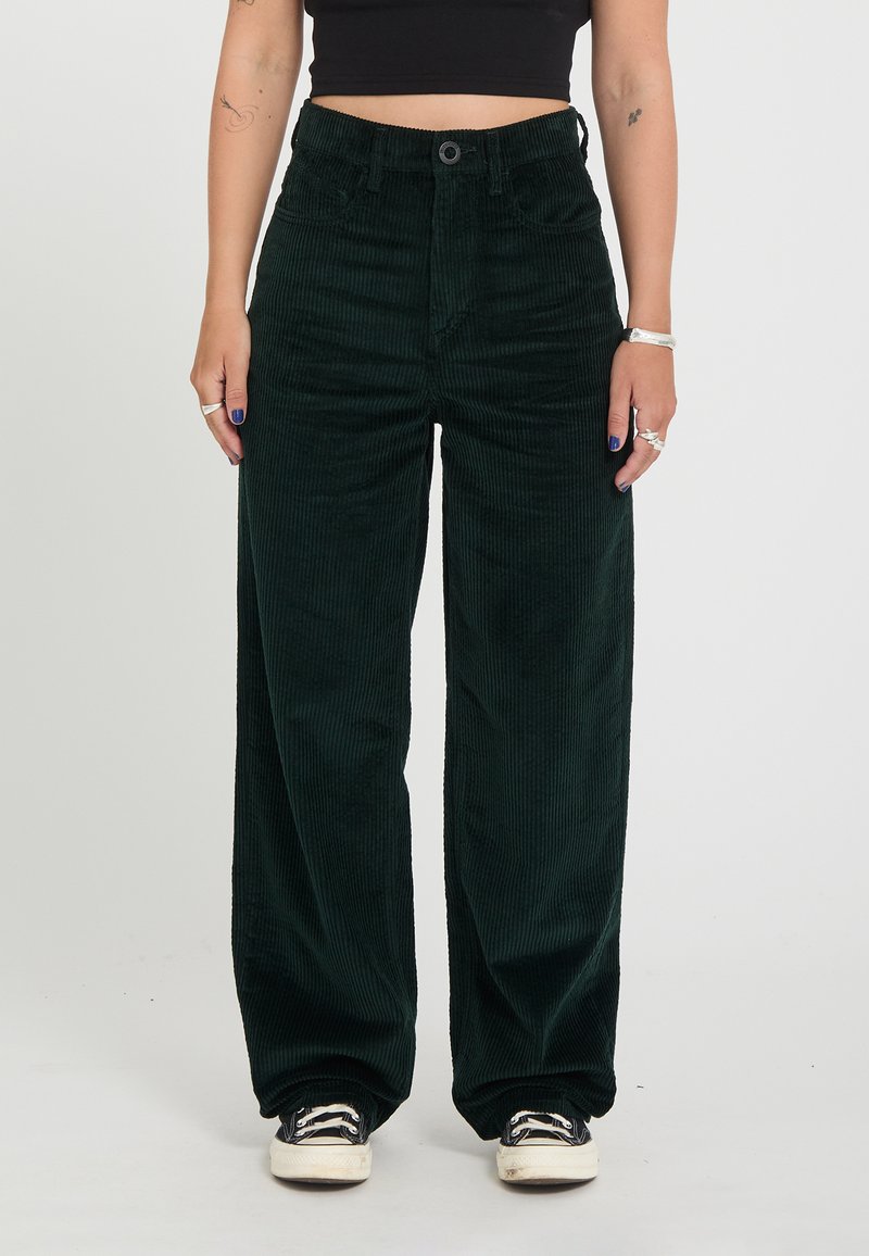 Personne portant un pantalon large en velours côtelé vert foncé taille haute, des baskets noires et blanches et un top court noir, debout devant un fond uni.