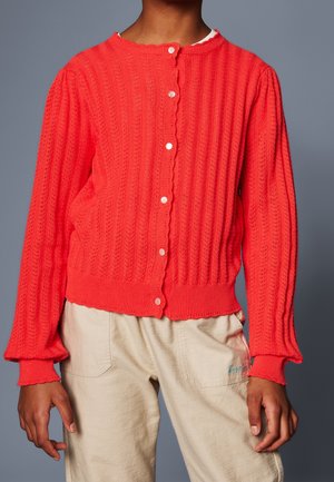 Personne portant un cardigan à boutons rouge vif texturé à manches longues et un pantalon beige, sur un fond gris uni.