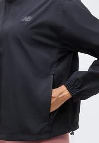 Sort New Balance jakke med lynlås foran og lynlåslomme i siden, hånd delvist inde i lommen, synlige elastiske manchetter.