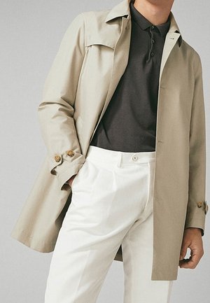 Beige trenchcoat met een gestructureerde kraag en houten knoopaccenten, gecombineerd met een donker polo shirt en witte geplisseerde broek.