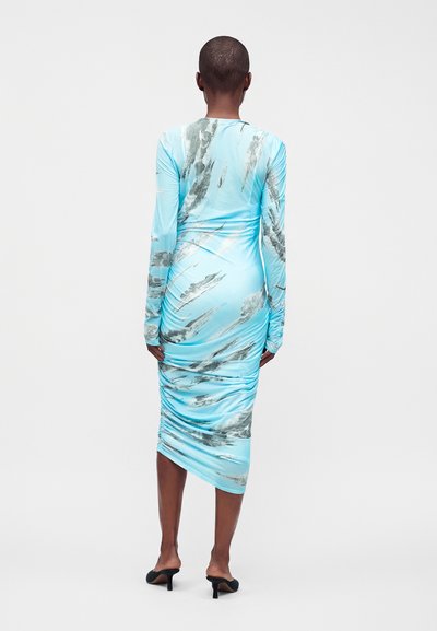 Personne debout, de dos, portant une robe bleu clair à manches longues froncée avec des motifs abstraits gris et des mules noires à talons.