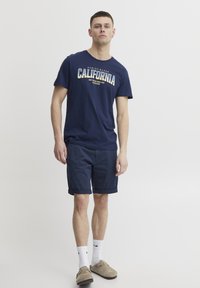 Jūras spēku kokvilnas T-krekls ar "California" grafiku, apvienots ar jūras spēku šortiem. Modelis nēsā baltas zeķes un bēšas slip-on kurpes. Vienkāršs dizains.