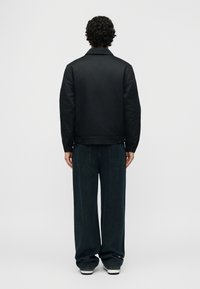 Άτομο με σγουρά μαλλιά, γυρισμένο με την πλάτη, φοράει μαύρο μπουφάν, χαλαρά σκουρόχρωμα τζιν και λευκά αθλητικά παπούτσια σε απλό φόντο.