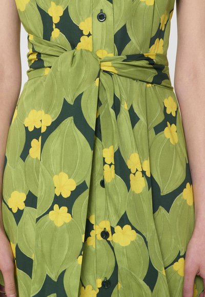 Gros plan d'une robe verte avec un grand motif de feuilles et de fleurs jaunes, dotée d'une ceinture à la taille et de boutons noirs sur le devant.