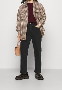 Camicia giacca oversize in motivo houndstooth marrone, jeans scuri, scarpe nere lucide e una piccola borsa beige. Outfit informale con texture miste.